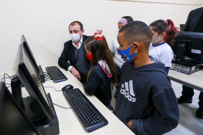 A foto mostra o governador Carlos Moisés sentado em frente a um computador, ele utiliza máscara e está acompanhado de cinco crianças, também de máscara. Cinco escolas da região devem receber laboratórios de informática, escolas de 13 municípios receberão equipamentos.