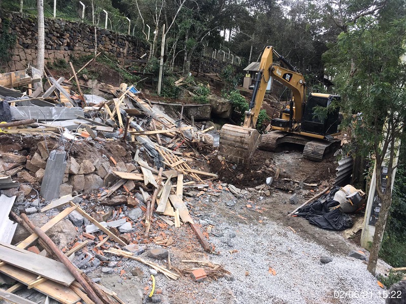 Uma escavadeira aparece no canto e ao lado, mais a frente, aparecem restos da demolição das obras irregulares em Florianópolis, com pedaços de pedra e madeiras