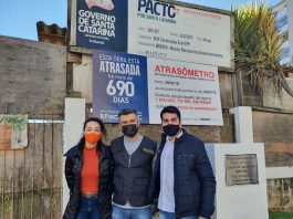 Deputado instala placa de “atrasômetro” em obra do colégio Aderbal Ramos da Silva políticos do novo usando máscara posam abraçados para foto em frente à obra e placa de "atrasômetro"
