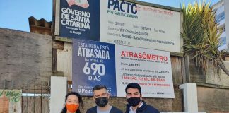 políticos do novo usando máscara posam abraçados para foto em frente à obra e placa de "atrasômetro"