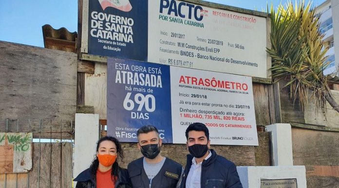 políticos do novo usando máscara posam abraçados para foto em frente à obra e placa de "atrasômetro"