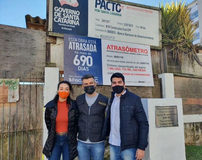 políticos do novo usando máscara posam abraçados para foto em frente à obra e placa de 