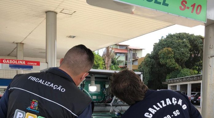 Duas pessoas de cabelo curto estão de costas olhando para uma bomba de um posto de combustível. Um veste um colete escuro do Procon de São José e outro veste uma camiseta preta em que se lê fiscalização imetro/sc. A operação transparência lacrou bombas irregulares durante a fiscalização em postos de combustíveis de são josé.