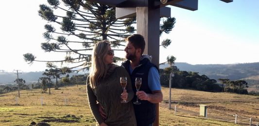 Roberta Zilli e Bruno Buske olham um para o outro em paisagem serrana segurando taça de vinho