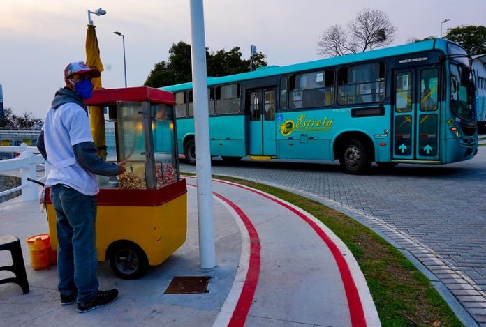 ônibus de transporte intermuicipal da empresa estrela saindo sda ponte herc´pilio luz e na calçada em primeiro plano pipoqueiro usando máscara com carrinho de pipoca