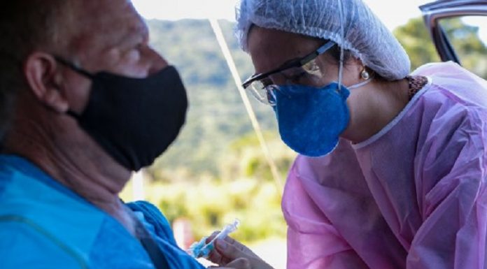 Um homem de azul e máscara preta sentado dentro de um carro é vacinado por uma profissional da saúde de máscara azul, touca branca e vestimenta rosa, que coloca a seringa no braço do senhor. Nesta semana, o estado bateu a marca de 3 milhões de doses de vacina aplicadas.