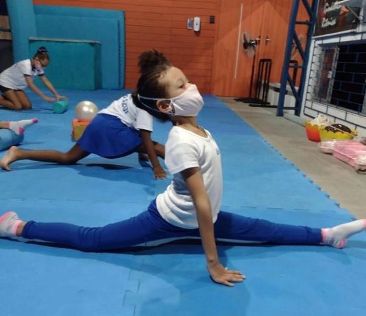 Menina de blusa branca, calça azul e máscara aparece de lado abrindo o espacate, em um tapete de azul claro e com outras crianças em volta. O time São José vai oferecer aulas de ginástica artística para crianças do município.