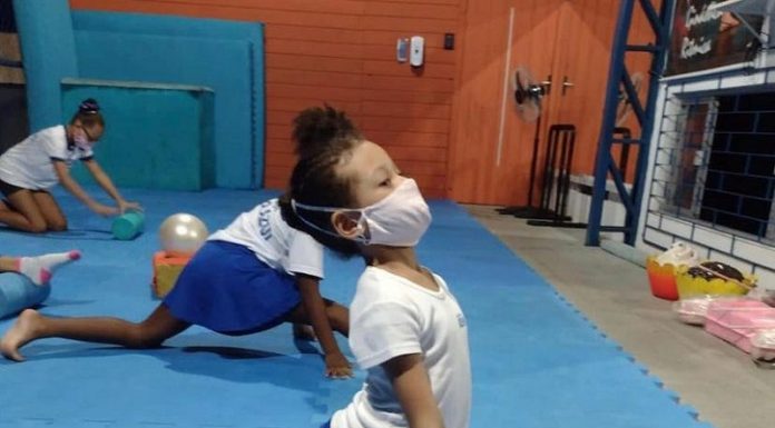 Menina de blusa branca, calça azul e máscara aparece de lado abrindo o espacate, em um tapete de azul claro e com outras crianças em volta. O time São José vai oferecer aulas de ginástica artística para crianças do município.