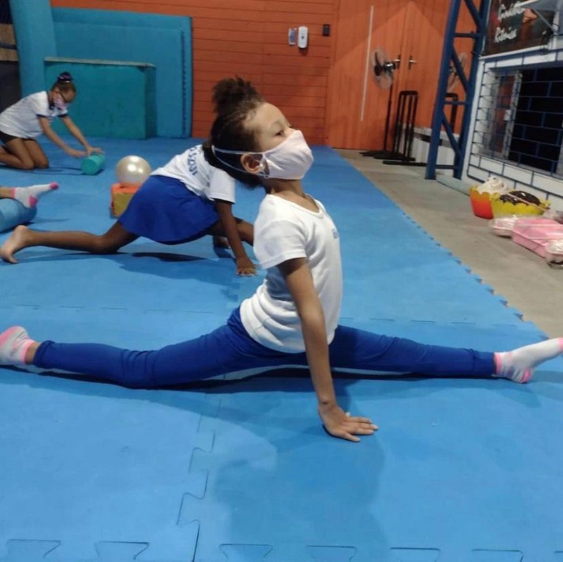 Menina de blusa branca, calça azul e máscara aparece de lado abrindo o espacate, em um tapete de azul claro e com outras crianças em volta. O time São José vai oferecer aulas de ginástica artística para crianças do município.