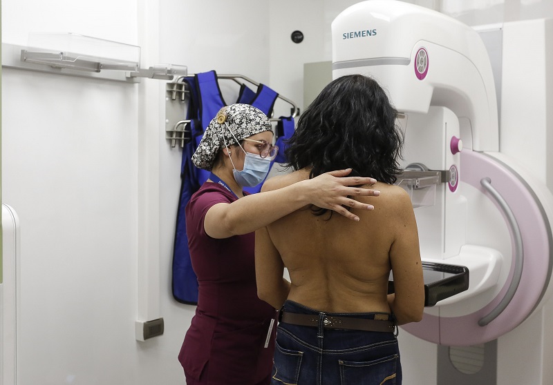 profissional de saúde orienta mulher sem camisa em frente a mamógrafo - Mulheres devem esperar para fazer mamografia após vacinação contra Covid