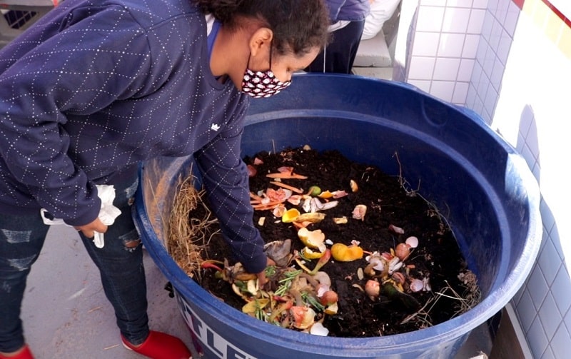 aluna usando máscara mexe em caixa d'água onde é feita a compostagem