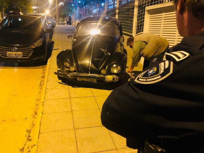 Parte de um guarda municipal aparece na foto de costas olhando para um fusca preto, que está ao centro da imagem em uma calçado. O veículo atropelou um motoboy em Florianópolis, no Itacorubi.