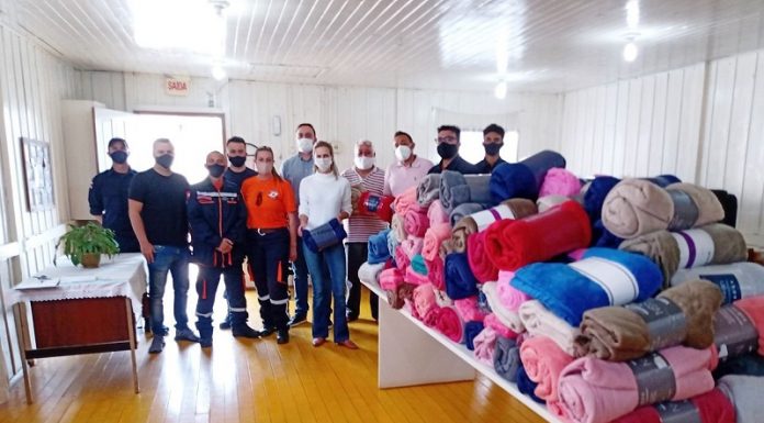 Um grupo de pessoas aparece lado a lado posando para a foto, ao lado, no canto direito, uma mesa com os cobertores doados para famílias da Frei Damiçao, em Palhoça.