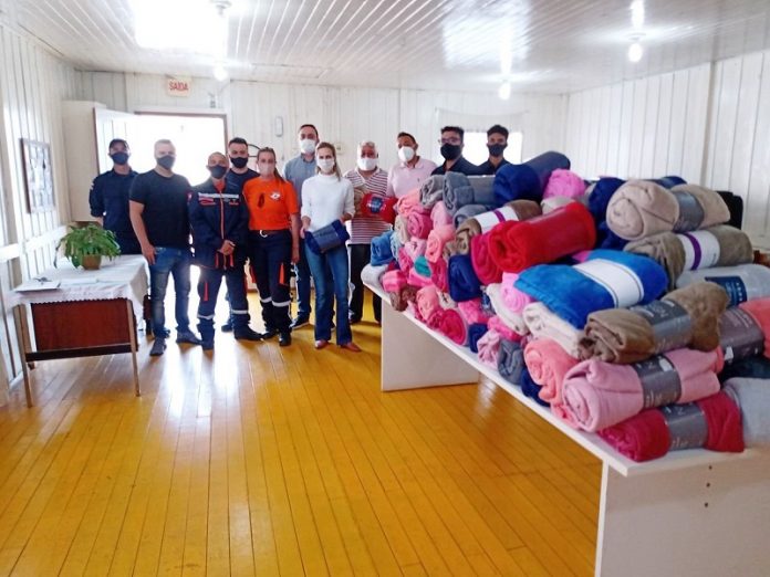 Um grupo de pessoas aparece lado a lado posando para a foto, ao lado, no canto direito, uma mesa com os cobertores doados para famílias da Frei Damiçao, em Palhoça.