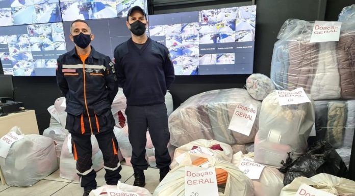 Dois homens com uniforme da defesa civil posam para a foto ao lado de sacos com doações da campanha cobertor solidário, que serão destinadas à comunidades de Palhoça.