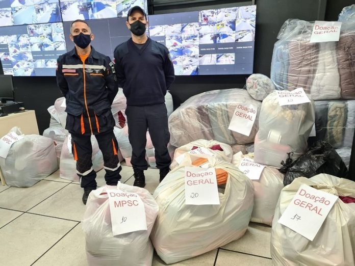 Dois homens com uniforme da defesa civil posam para a foto ao lado de sacos com doações da campanha cobertor solidário, que serão destinadas à comunidades de Palhoça.