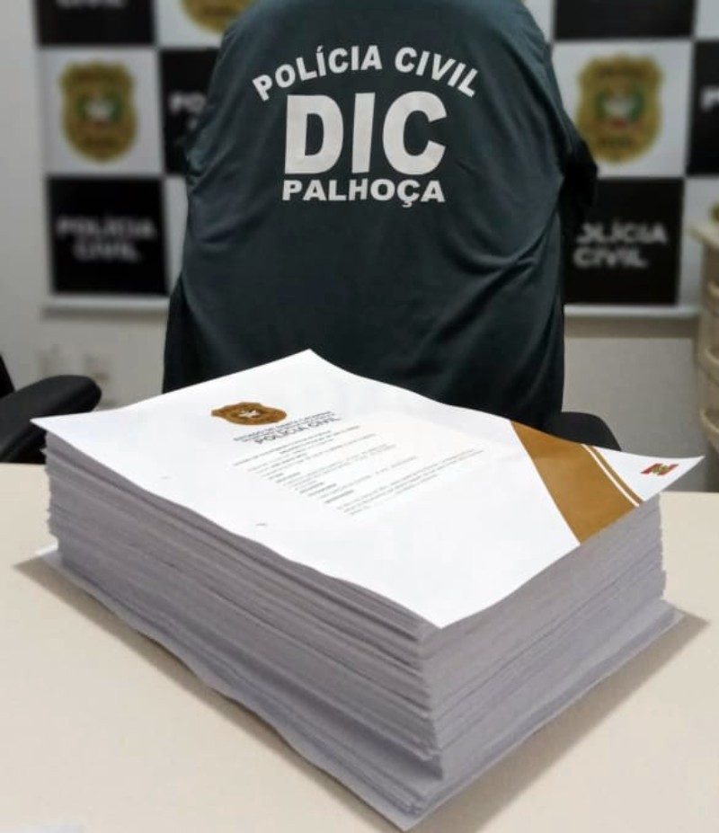 documento do inquérito com centenas de páginas sobre mesa com logo da dic e polícia civil ao fundo - 125 pessoas em Palhoça são acusadas de integrar facção criminosa