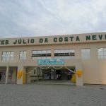Entrada da escola, identificada como EEB Júlio da Costa Neves, na Costeira, em Florianópolis. A estrutura tem cor amarelo claro e na entrada há um cartaz de bem vindo.