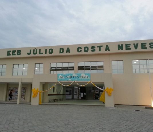 Entrada da escola, identificada como EEB Júlio da Costa Neves, na Costeira, em Florianópolis. A estrutura tem cor amarelo claro e na entrada há um cartaz de bem vindo.