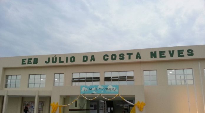 Entrada da escola, identificada como EEB Júlio da Costa Neves, na Costeira, em Florianópolis. A estrutura tem cor amarelo claro e na entrada há um cartaz de bem vindo.