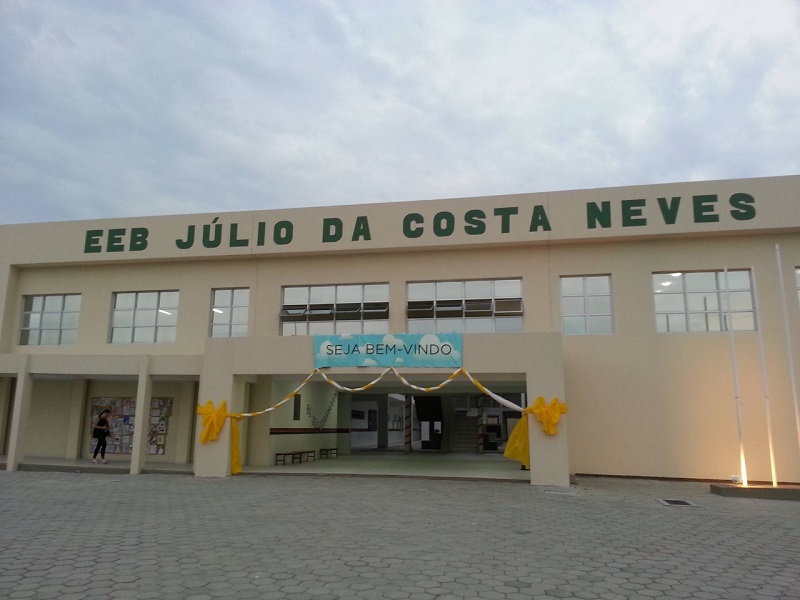 Entrada da escola, identificada como EEB Júlio da Costa Neves, na Costeira, em Florianópolis. A estrutura tem cor amarelo claro e na entrada há um cartaz de bem vindo.