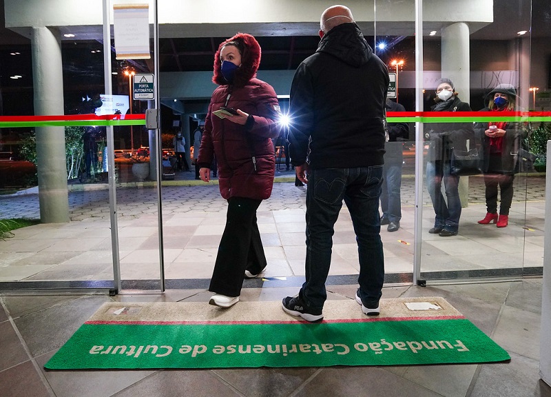 Na entrado do CIC, uma porta de vidro e um tapete verde com branco sinalizado com "fundação catarinense de cultura". Um homem de agasalho aparece de costas e uma mulher aparece caminhando para a estrado, do lado de fora outras pessoas se aproximam da porta. Santa Catarina teve nesta semana seu primeiro evento-teste com pessoas imunizadas contra a covid-19.