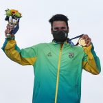 Ítalo Ferreira tem cabelos escuros e curtos, usa uma máscara preta e um agasalho verde e amarelo do Brasil, ele segura com a mão a medalha de ouro, conquistada no surfe, enroscada no pescoço e na outra mão levanta as flores entregues junto com a medalha nas olimpíadas de tóquio.