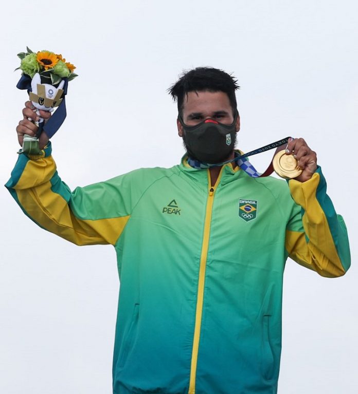 Ítalo Ferreira tem cabelos escuros e curtos, usa uma máscara preta e um agasalho verde e amarelo do Brasil, ele segura com a mão a medalha de ouro, conquistada no surfe, enroscada no pescoço e na outra mão levanta as flores entregues junto com a medalha nas olimpíadas de tóquio.