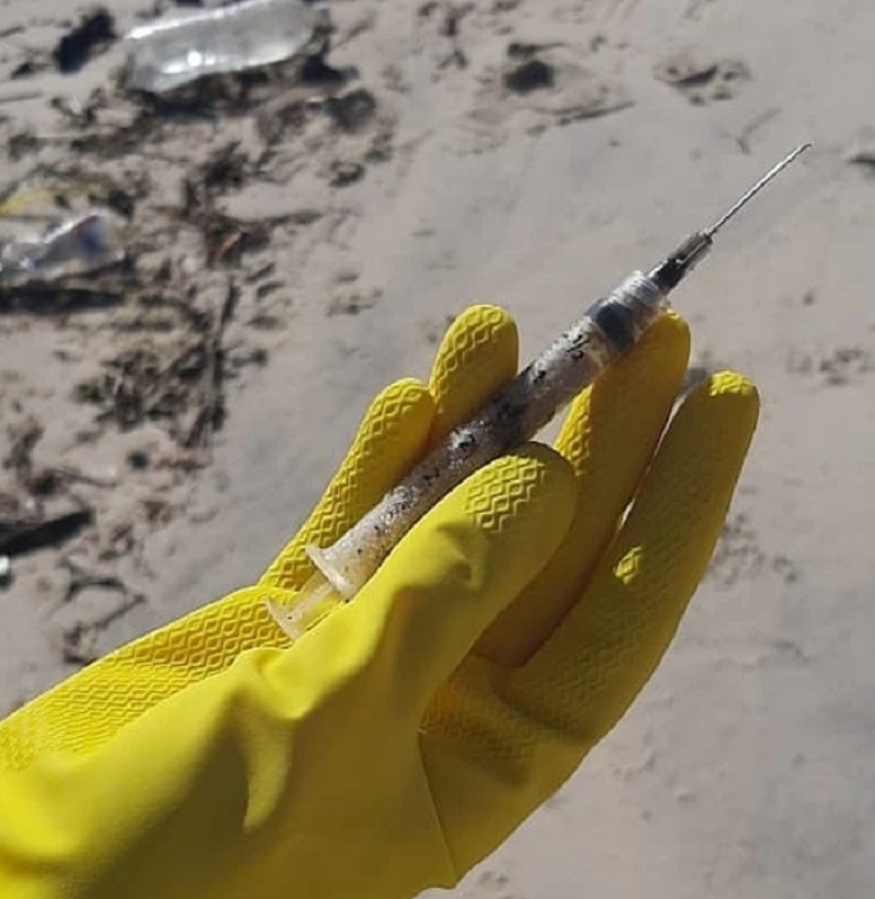 Uma mão com luva amarela segura uma seringa com agulha. Esse e outros resíduos foram recolhidos pelo mutirão da Palhoça menos Lixo.