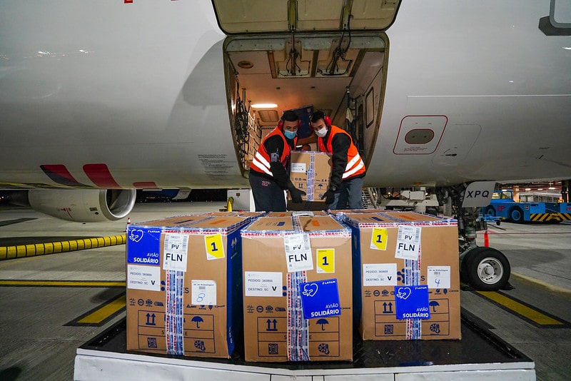 dois homens usando colete e protetores de ouvido retiram caixas de papelão de bagaeiro de avião - Lote insuficiente não amplia vacinação de Covid na Grande Florianópolis