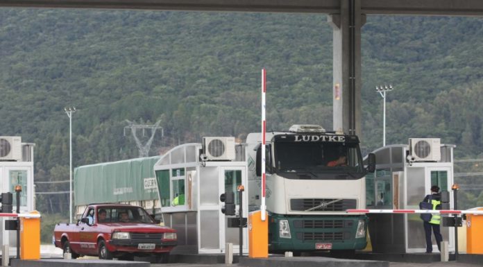 Um caminhão branco com preto está parado em um ponto de pedágio, ao lado um carro vermelho também está pagando a tarifa. Um projeto de lei foi aprovado em santa catarina, autorizando o pagamento com cartão das tarifas de pedágio, mas a concessionária contesta.