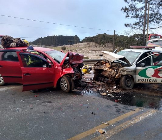 A foto mostra um veículo vermelho de frente para uma viatura da polícia militar, os dois veículos têm a parte da frente destruídas após uma colisão frontal, que deixou um policial morto e outros feridos em ANita Garibaldi. A pista tinha gelo no momento do acidente.