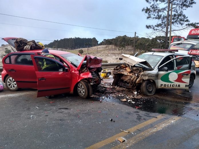 A foto mostra um veículo vermelho de frente para uma viatura da polícia militar, os dois veículos têm a parte da frente destruídas após uma colisão frontal, que deixou um policial morto e outros feridos em ANita Garibaldi. A pista tinha gelo no momento do acidente.