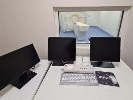 Em uma mesa branca, três telas pretas e ao fundo uma sala de tomografia. O hospital universitário da UFSC adquiriu um novo tomógrafo para exames, atendendo a população.