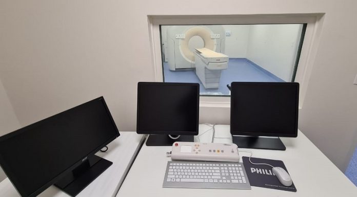 Em uma mesa branca, três telas pretas e ao fundo uma sala de tomografia. O hospital universitário da UFSC adquiriu um novo tomógrafo para exames, atendendo a população.