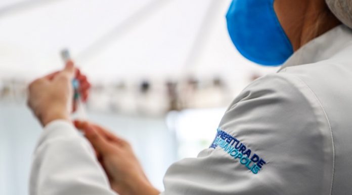 Vacinação contra a gripe no público geral inicia em Florianópolis nesta terça A foto foca no ombro do profissional de saúde, na parte do colete em que se lê prefeitura de Florianópolis. Este profissional veste além do colete azul, uma máscara azul e manipula uma seringa de vacina. A foto ilustra a vacinação contra a gripe, que em Florianópolis começa a atender o público geral.