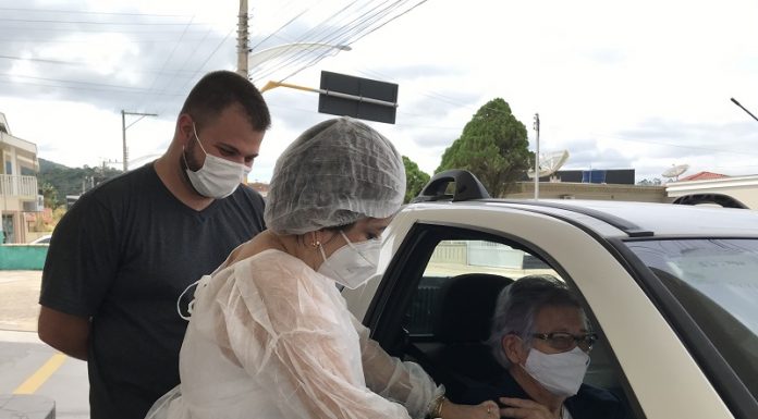 Uma profissional de saúde com vestimenta brancas aplica a vacina da covid-19 em um senhor dentro de um veículo, em Nova Trento, ao lado do carro, do lado de fora, um rapaz observa. O município de Nova Trento abriu nesta semana o cadastro para xepa da vacina contra a covid-19.