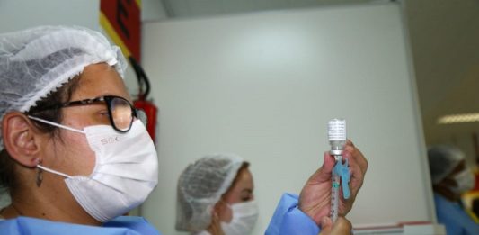 Xepa da vacina é liberada para adolescentes com deficiência ou comorbidades Uma profissional de saúde com vestimentas azuis e máscara e touca branca, além de ´pculos de grau, segura a sua frente um frasco e uma seringa de vacina. A foto ilustra a notícia da liberação do uso de xepa da vacina para adolescentes comd eficiências ou comorbidades em SC.