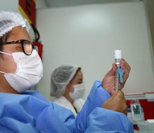 Uma profissional de saúde com vestimentas azuis e máscara e touca branca, além de ´pculos de grau, segura a sua frente um frasco e uma seringa de vacina. A foto ilustra a notícia da liberação do uso de xepa da vacina para adolescentes comd eficiências ou comorbidades em SC.