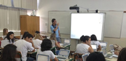 Uma sala de aula com um professor a frente olhando para um telão e observado por estudantes nas carteiras com computadores. O novo ensino médio em SC entra em vigor em 2022 nas escolas estaduais.