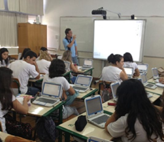 Uma sala de aula com um professor a frente olhando para um telão e observado por estudantes nas carteiras com computadores. O novo ensino médio em SC entra em vigor em 2022 nas escolas estaduais.