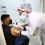Um homem de cabelo escuro veste camiseta e máscara pretas, ele está sentado e recebe a vacina contra a covid no braço, aplicada por uma profissional da saúde com vestimentas brancas. Neste sábado, Florianópolis vai ampliar a vacinação contra a Covid-19 para pessoas de 26 anos.