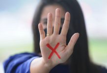 Mulher branca de cabelo comprido e escuro mostra a palma da mão para a foto com um x vermelho desenhado na palma, o sinal simboliza a denúncia contra a violência doméstica contra a mulher, contemplada na lei maria da penha
