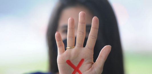Mulher branca de cabelo comprido e escuro mostra a palma da mão para a foto com um x vermelho desenhado na palma, o sinal simboliza a denúncia contra a violência doméstica contra a mulher, contemplada na lei maria da penha