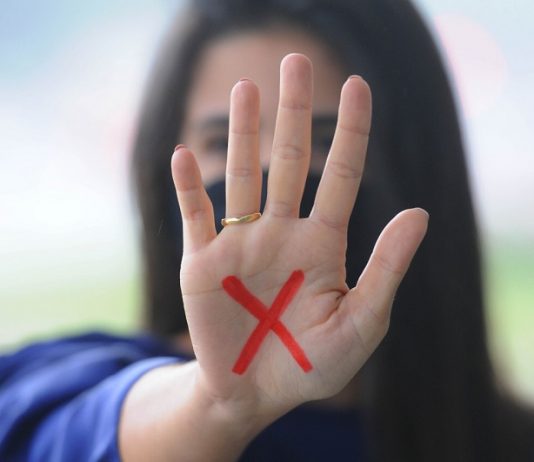 Lei Maria da Penha completa 15 anos entre avanços e persistência da violência Mulher branca de cabelo comprido e escuro mostra a palma da mão para a foto com um x vermelho desenhado na palma, o sinal simboliza a denúncia contra a violência doméstica contra a mulher, contemplada na lei maria da penha