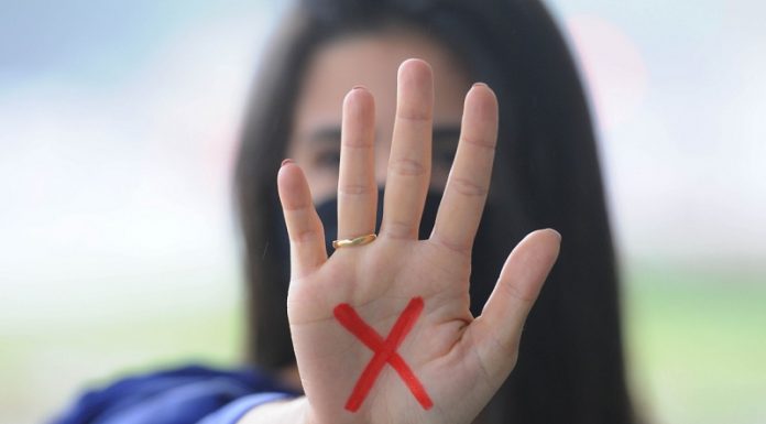 Mulher branca de cabelo comprido e escuro mostra a palma da mão para a foto com um x vermelho desenhado na palma, o sinal simboliza a denúncia contra a violência doméstica contra a mulher, contemplada na lei maria da penha