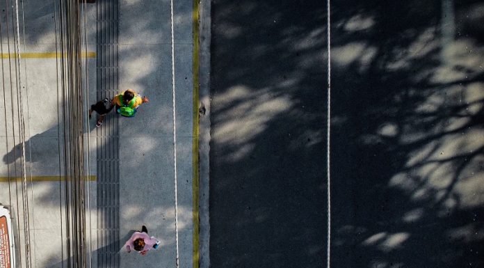 Vista de cima mostra uma rua cinza com linha branca à direita, à esquerda uma calçada cinza com dois pedestres em movimentos. A imagem ilustra a notícia de que Biguaçu vai notificar proprietários de imóveis para adequação ou implantação de calçadas.