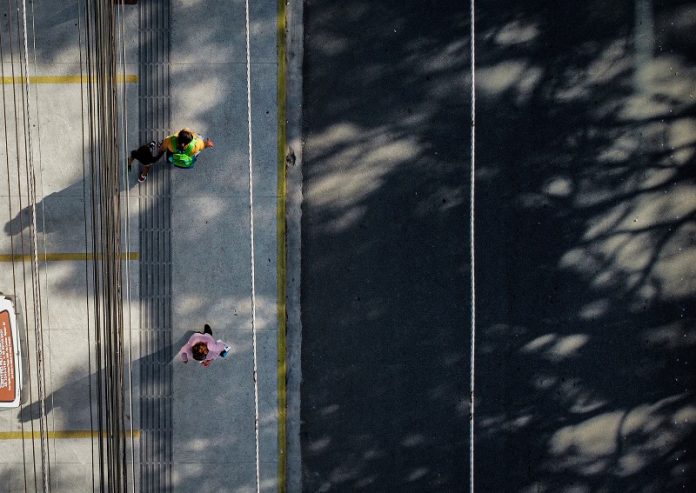 Vista de cima mostra uma rua cinza com linha branca à direita, à esquerda uma calçada cinza com dois pedestres em movimentos. A imagem ilustra a notícia de que Biguaçu vai notificar proprietários de imóveis para adequação ou implantação de calçadas.