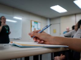 Edital publicado para concurso de ACTs em Santa Catarina Edital publicado para concurso de ACTs em Santa Catarina - close em mão segurando caneta sobre caderno em sala de aula e professora em pé ao fundo dando aula