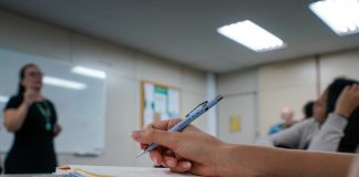 Edital publicado para concurso de ACTs em Santa Catarina - close em mão segurando caneta sobre caderno em sala de aula e professora em pé ao fundo dando aula